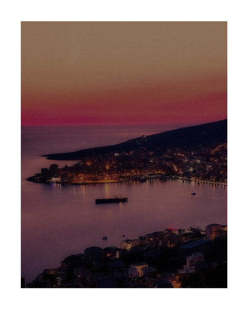 Vista costeira ao entardecer no verão europeu, com céu em tons rosados, mar calmo e luzes da cidade ao redor da baía