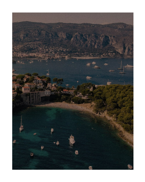 Vista aérea de enseada no verão europeu, com barcos ancorados, mar azul-turquesa, vegetação e cidade costeira ao fundo