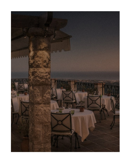 Restaurante com varanda ao ar livre no verão europeu, com mesas postas, vista para o mar e atmosfera elegante ao entardecer