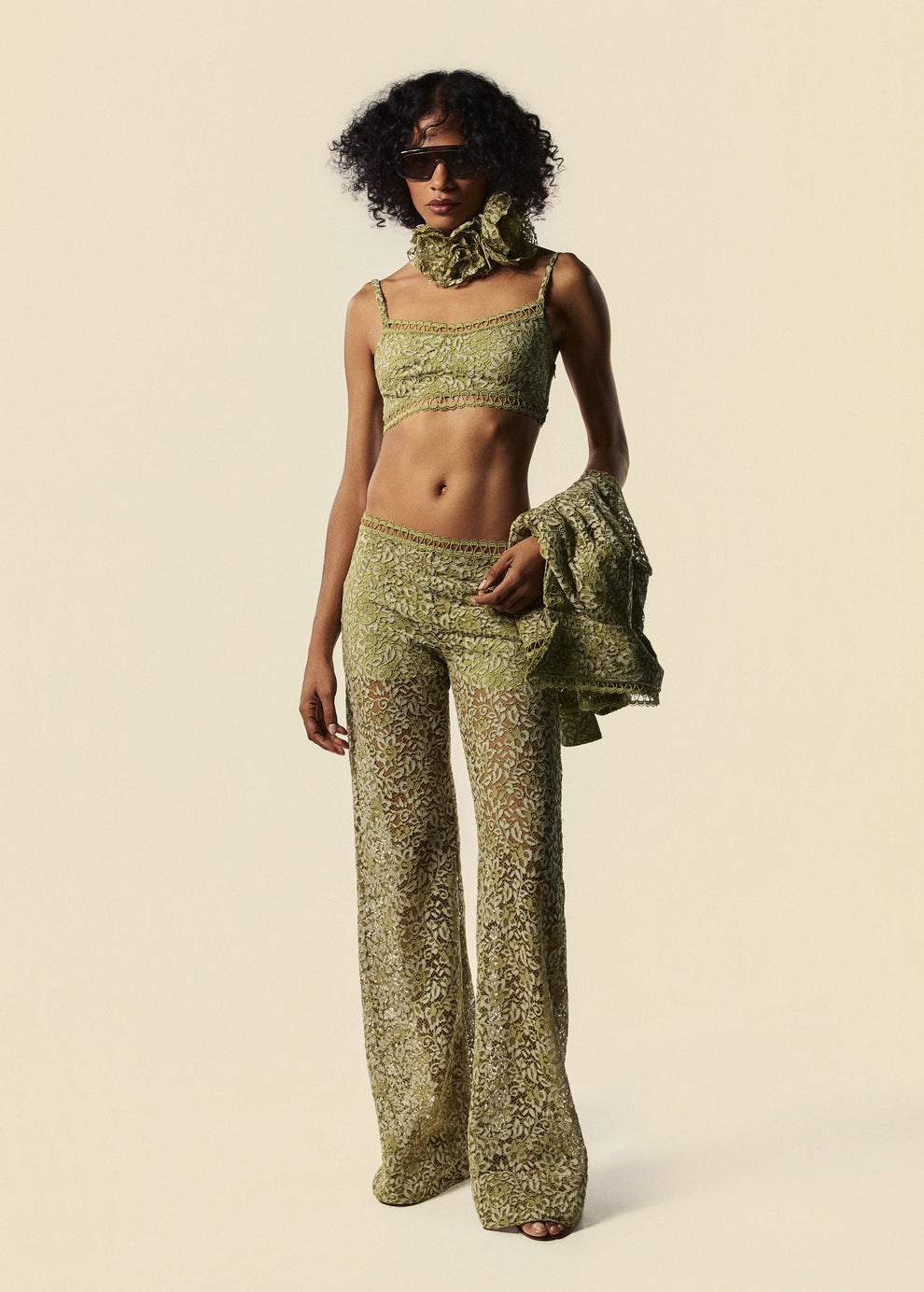 Modelo segura camisa feminina, mostrando o top curto e calça flare de renda com colar de flores em verde do lançamento da La Gea.
