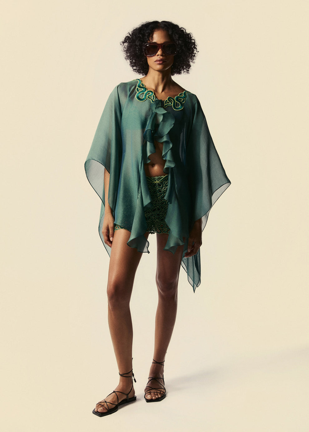 Modelo combina kaftan curto verde Destino com shorts mini Trilhas e rasteirinha de tiras pretas e óculos de sol.