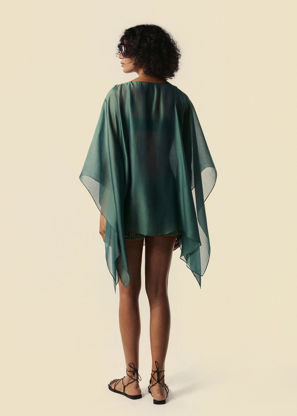 Modelo de costas com o kaftan curto verde Destino com mangas amplas e shorts mini Trilhas com rasteirinha de tiras na cor preta.