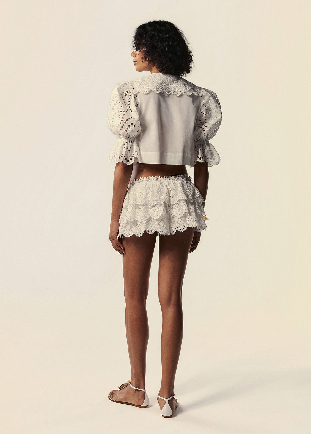 Modelo de costas com detalhes do shorts mini e camisa cropped em laise branco da família Lúmina da La Gea.