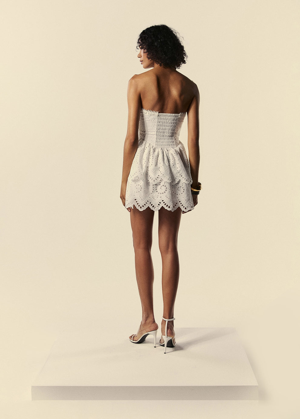 Modelo de costas com o vestido curto branco em laise com babados e lastex estilo tomara que caia da família Lumina.