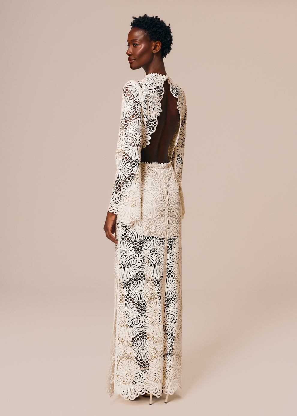 Modelo de costas usa vestido longo com manga longa e fenda lateral em renda off White Cerâmica La Gea