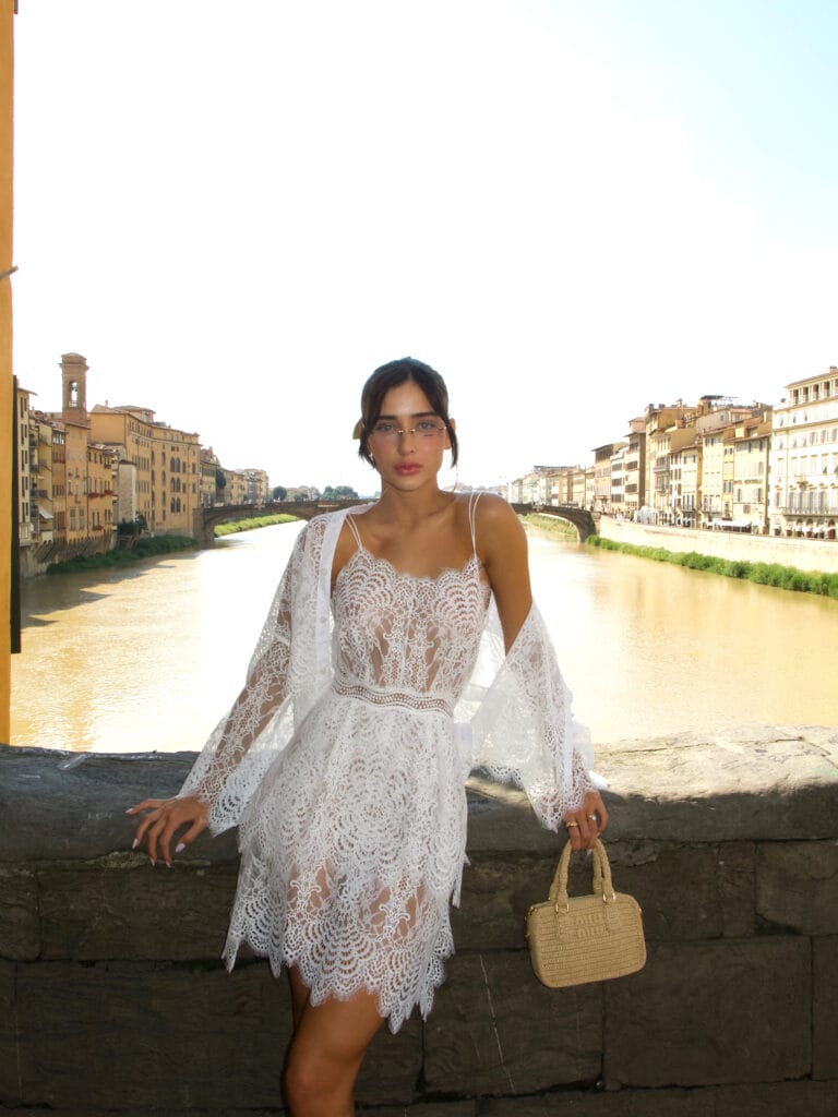 Mulher usando um vestido rendado branco e uma bolsa pequena, posando em frente a um rio.