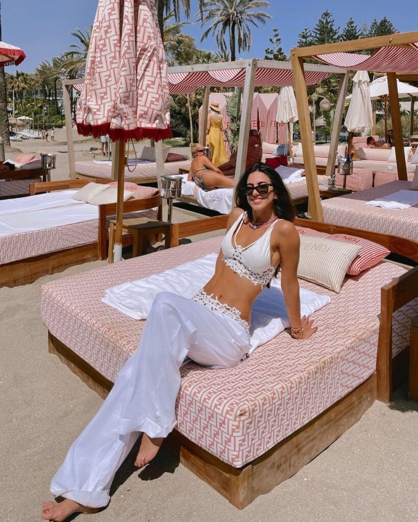 Mulher de calça pantalona branca e top branco posando em cama de sol de um resort wear.