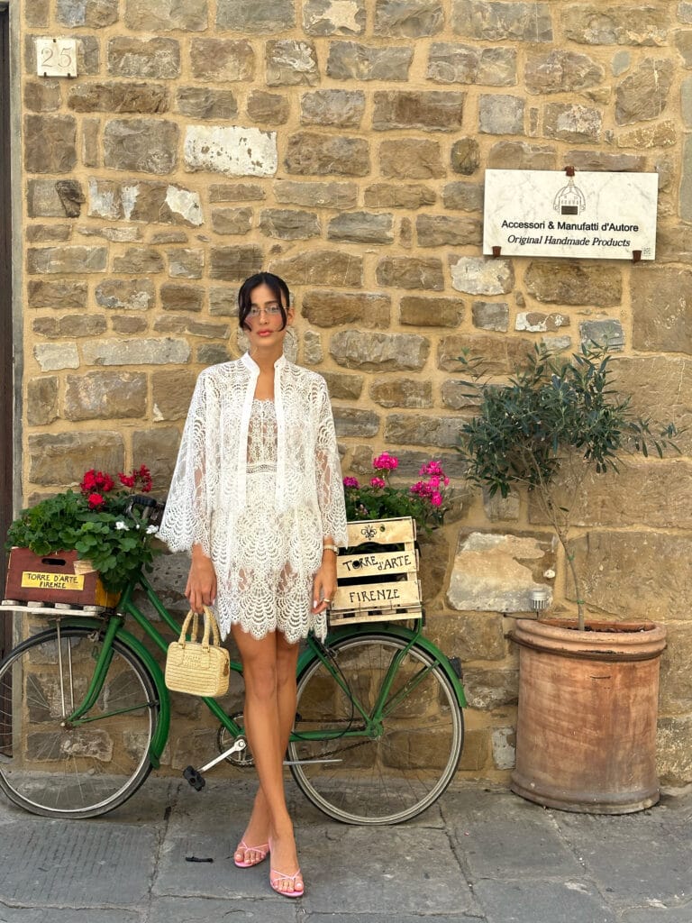Mulher usando um vestido rendado branco e uma bolsa pequena, posando em frente a um muro de pedras e uma bicicleta verde