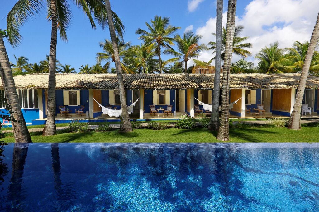  Imagem com piscina à frente com pé de coqueiro e casas no fundo da imagem com varanda